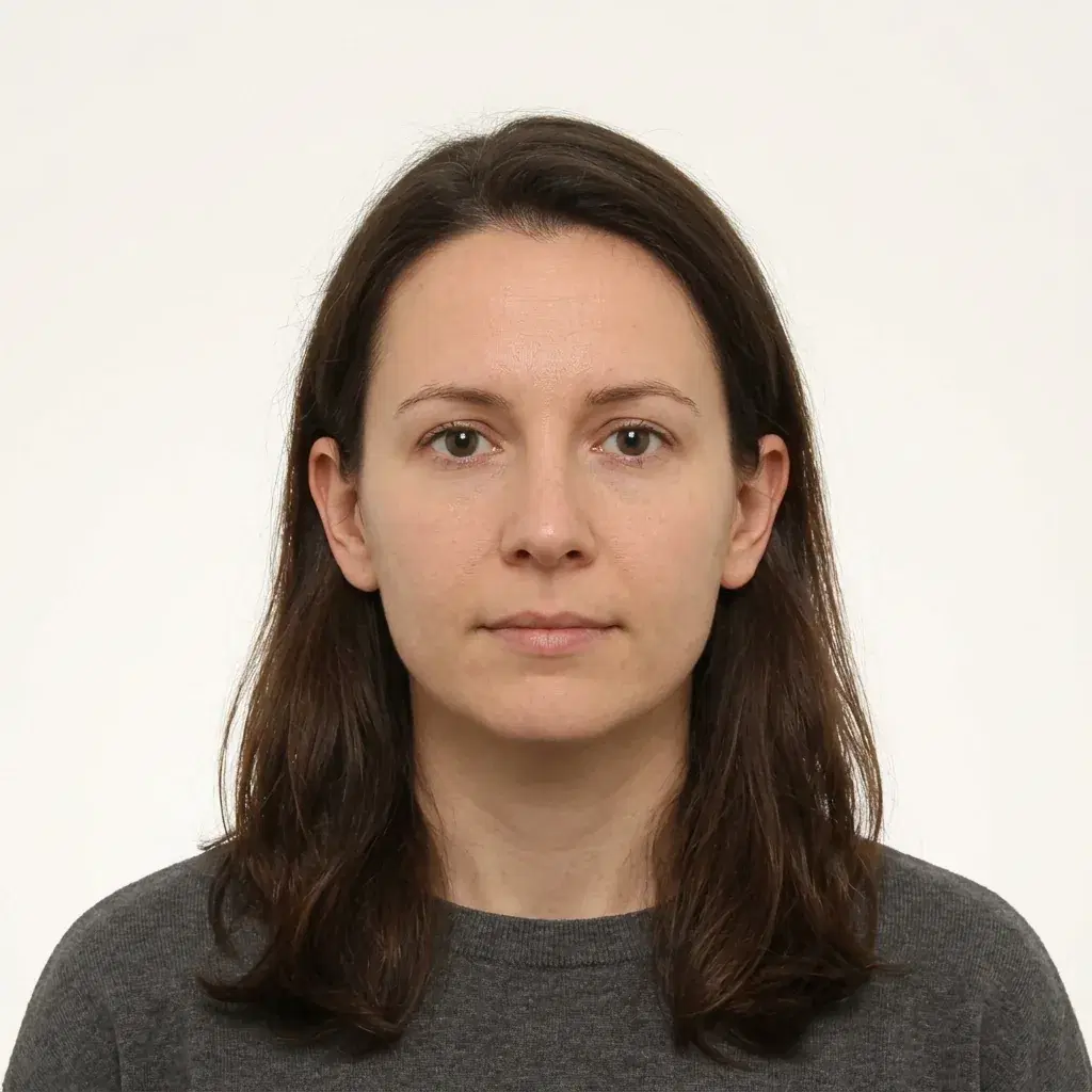 Passport-style portrait of a woman with dark hair in a charcoal top, facing the camera on a plain off-white background